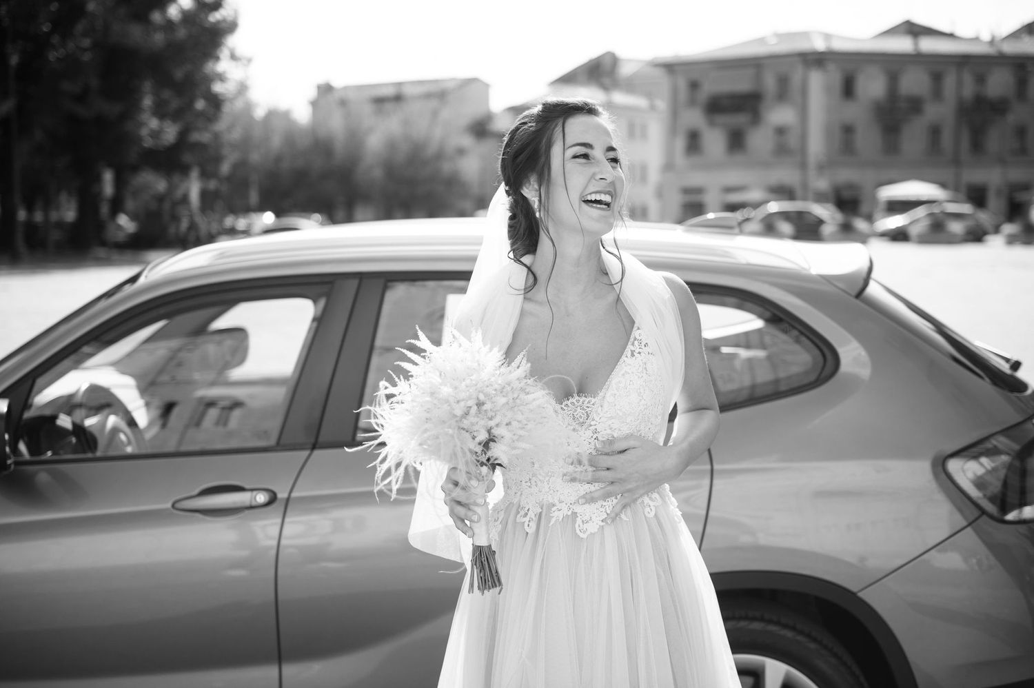 A joyful bride laughing while holding a white bouquet next to a parked car.