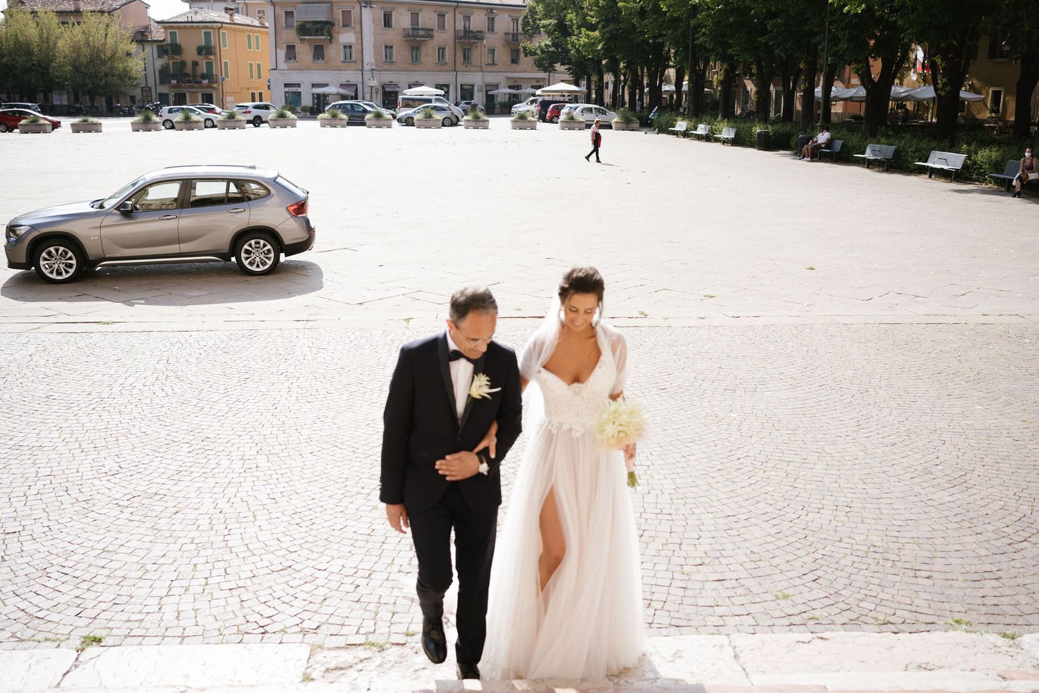 A wedding couple walking together across a sunny plaza with parked cars in the background.