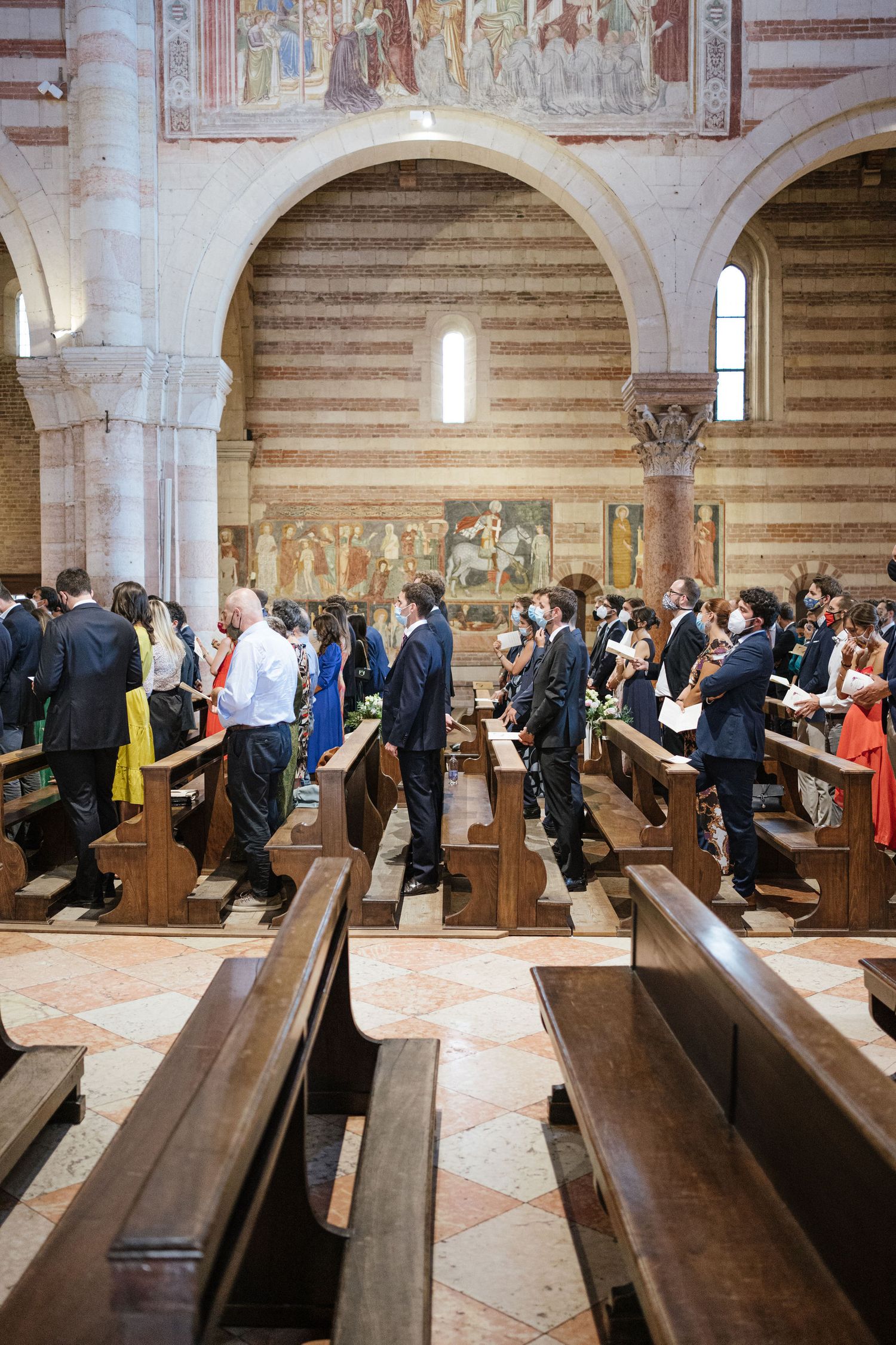 Wedding guests standing in wooden pews inside a church with striped walls and religious frescoes.