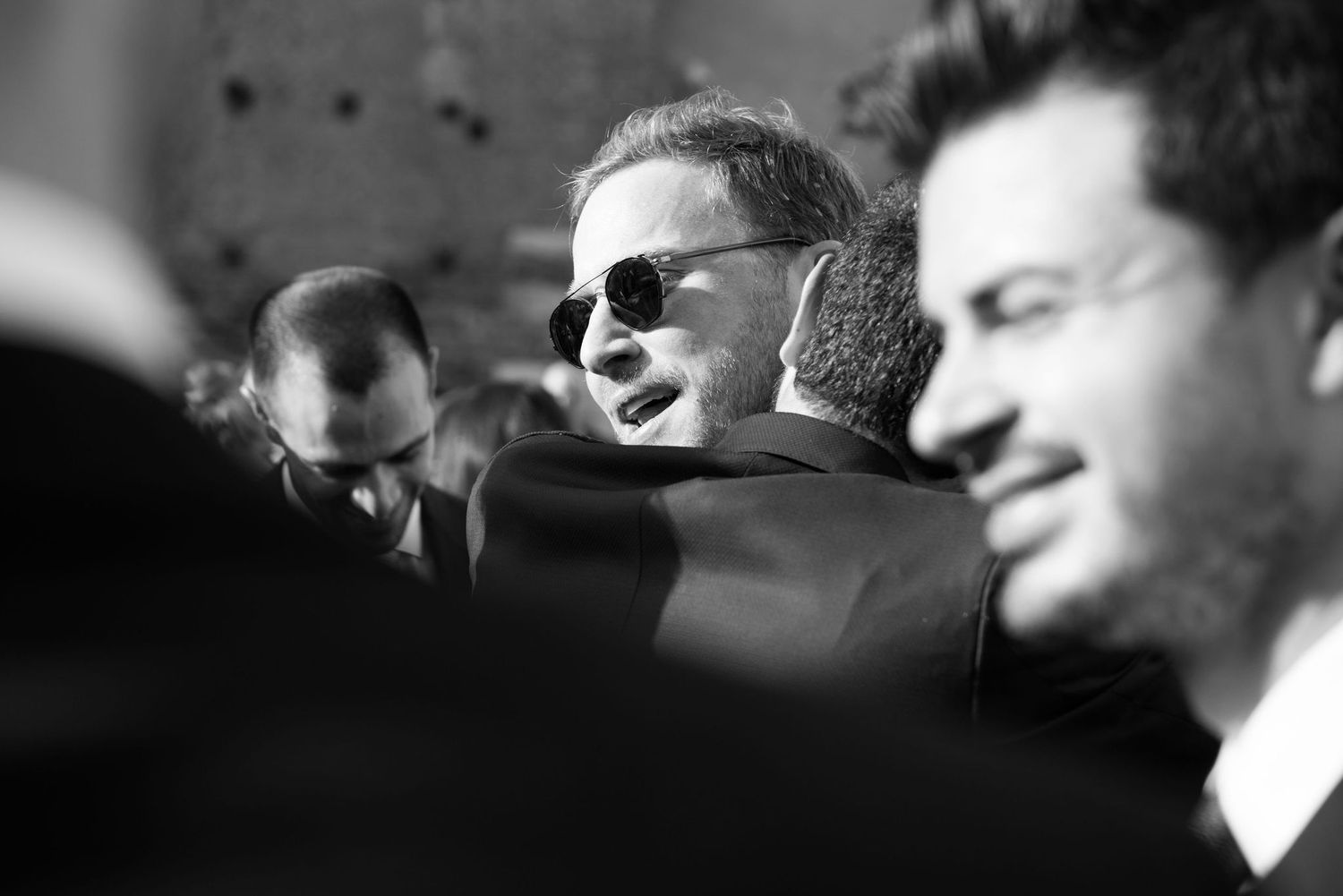 Black and white candid photo showing a crowd gathered outdoors with one person wearing sunglasses in focus.