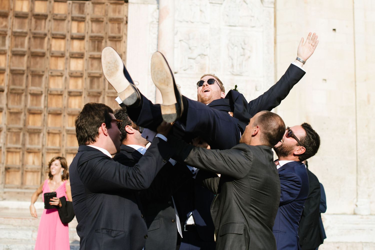 Groomsmen in black suits tossing someone in the air during wedding celebrations outside a church.