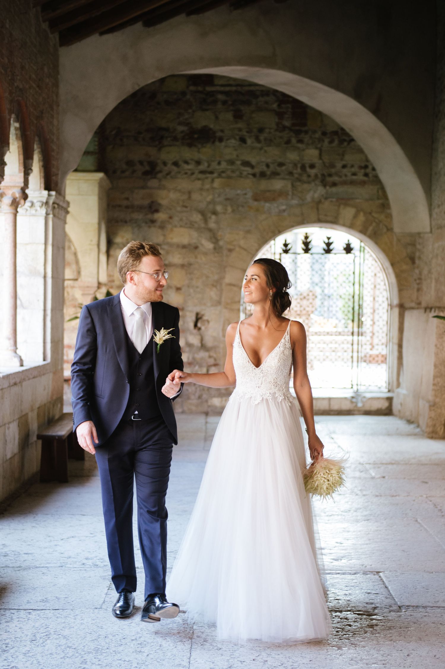 Elegant wedding couple walking through a historic stone archway corridor.