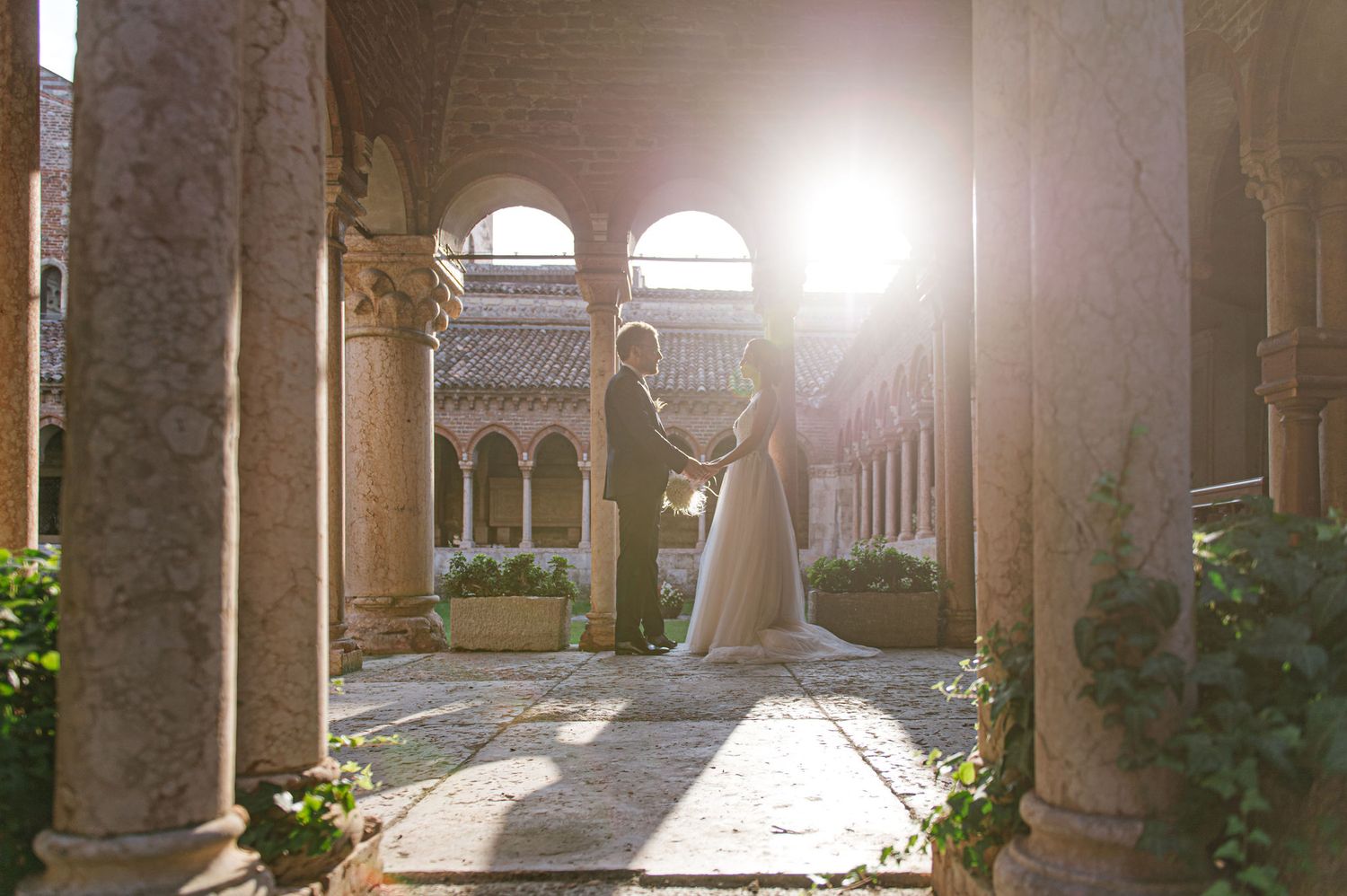 Wedding couple embraces in sunlit stone courtyard with elegant archways and columns.