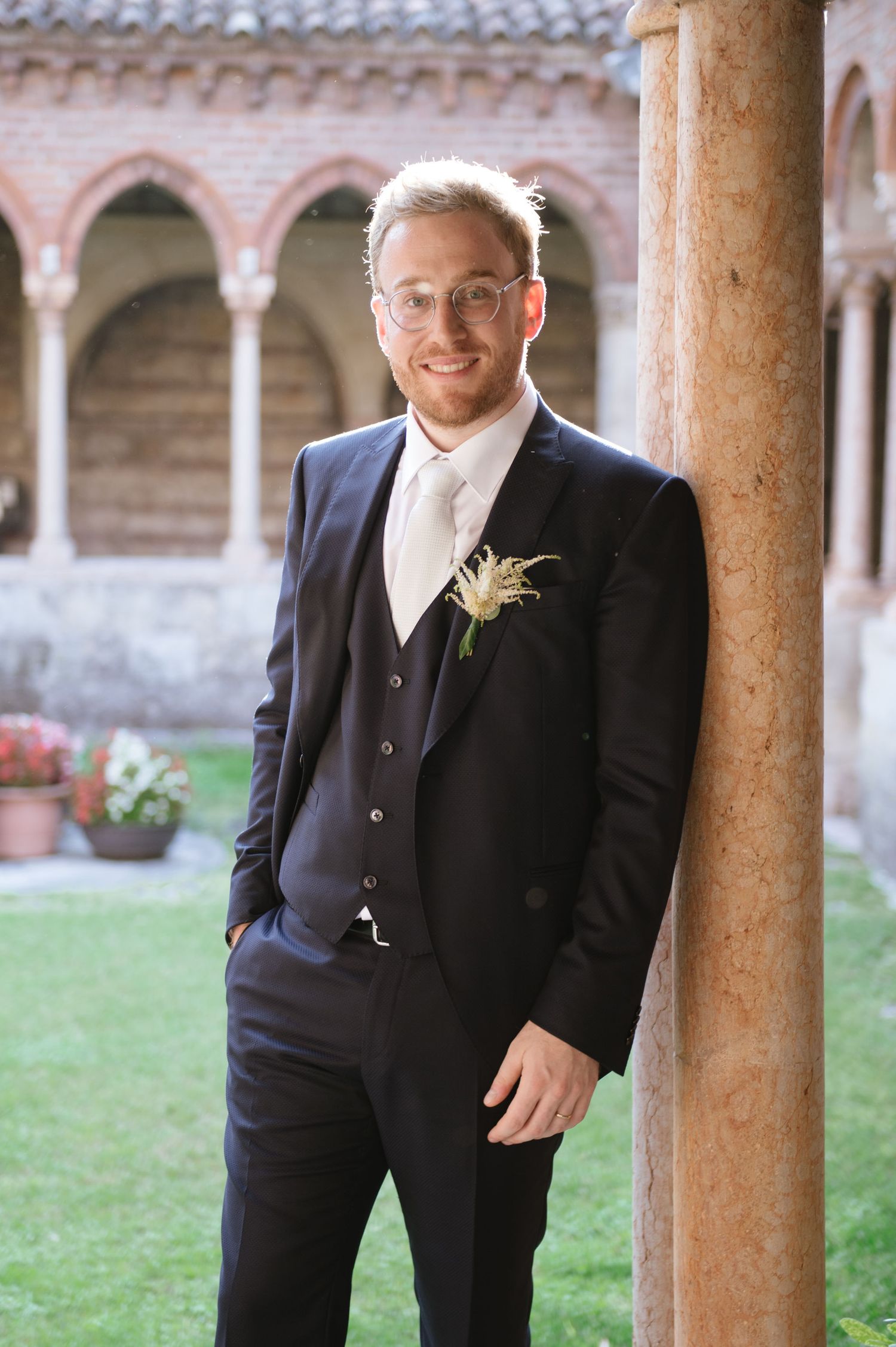 Groom in navy suit with boutonniere poses against stone archway in garden courtyard.