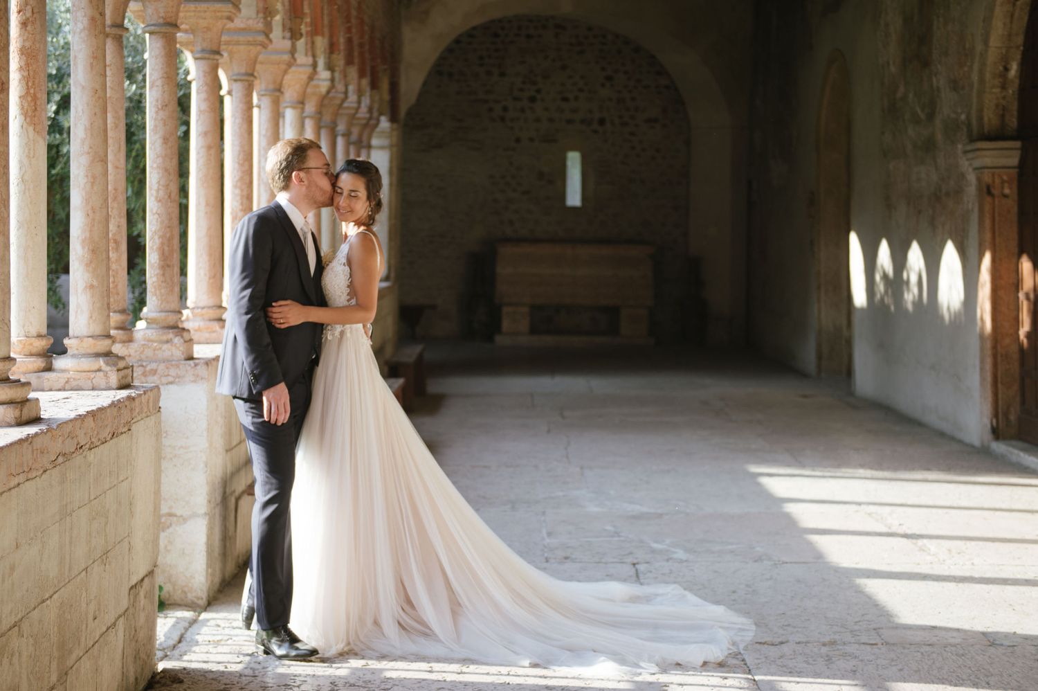 Couple stands in sunlit stone corridor with flowing white wedding dress train.