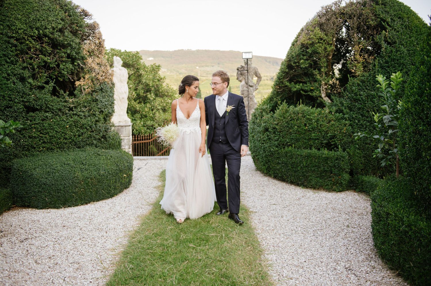Couple poses on garden path surrounded by manicured hedges and greenery.