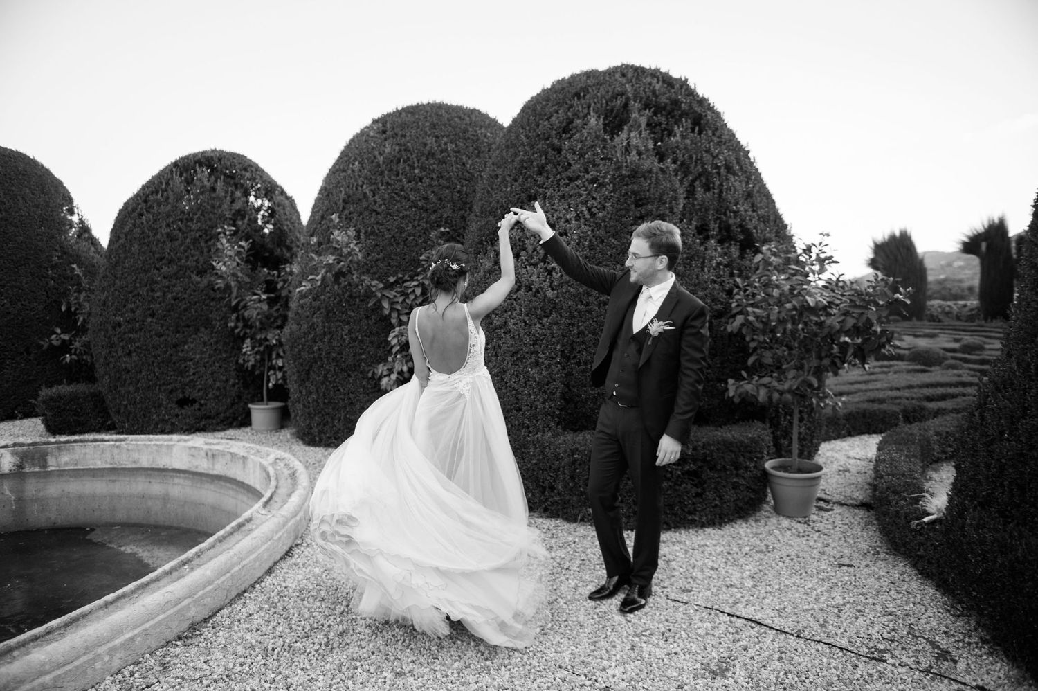 Black and white photo of dancing couple in formal wear by manicured garden topiary.