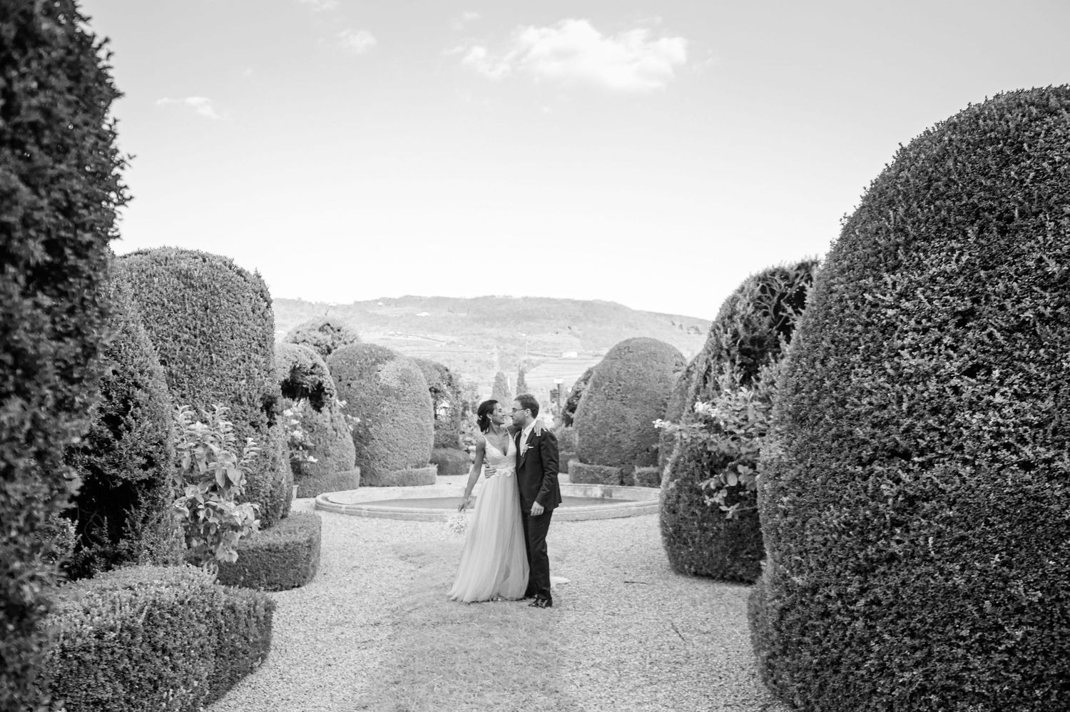 Black and white wedding photo among sculptured garden hedges with distant hills.