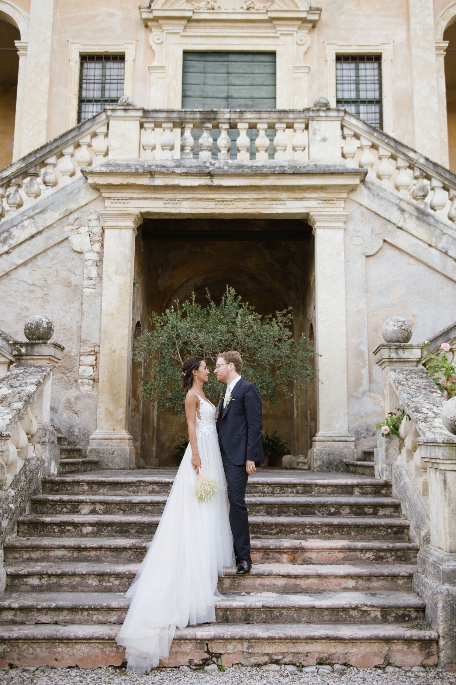 A romantic wedding couple stands on ornate stone steps of an Italian villa with classic architecture.
