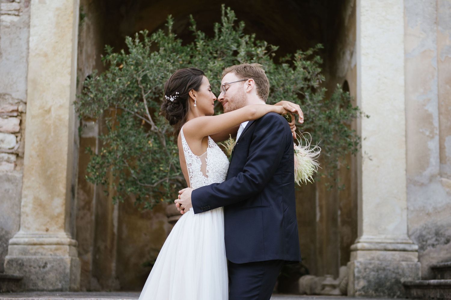 Couple shares romantic moment under stone archway with flowering greenery.