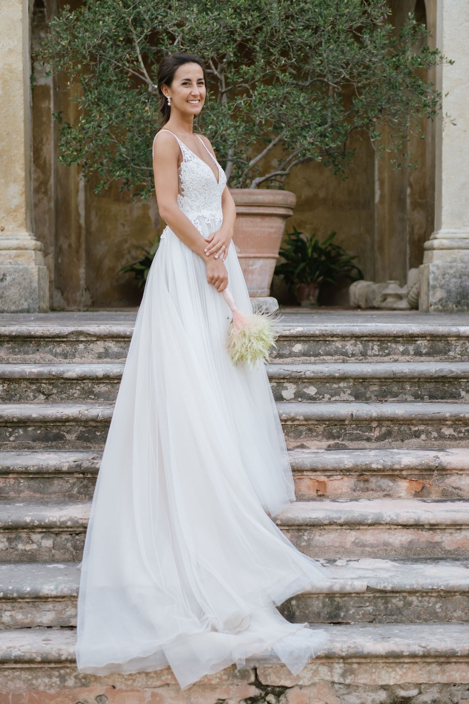 A bride in a flowing white gown stands elegantly on historic stone steps with greenery behind.