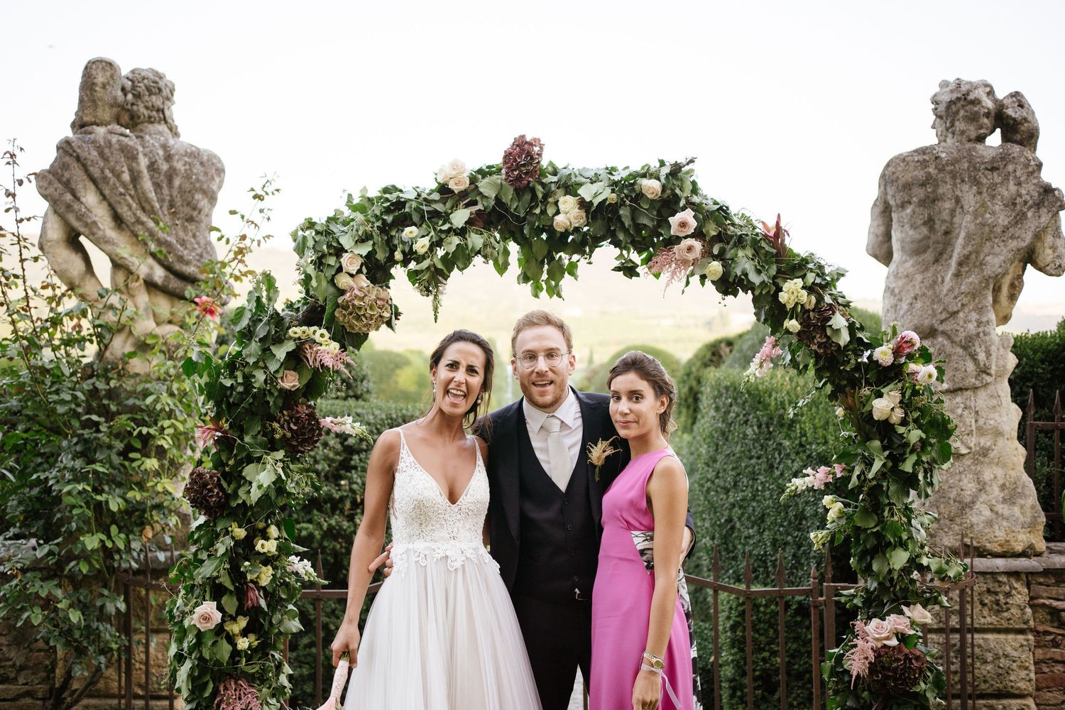 A wedding party poses under a lush floral arch between stone statues in a garden setting.