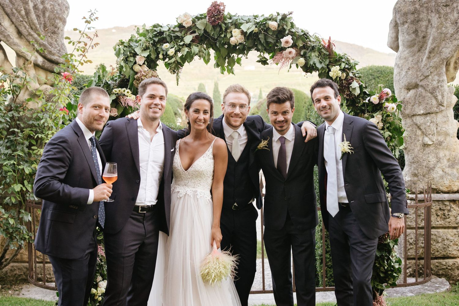 Wedding party members gather under a floral arch for a celebratory group photo.