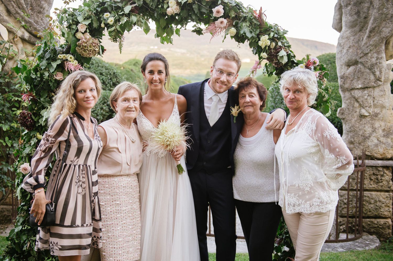 Wedding group photo under a decorative floral arch in an elegant garden setting.