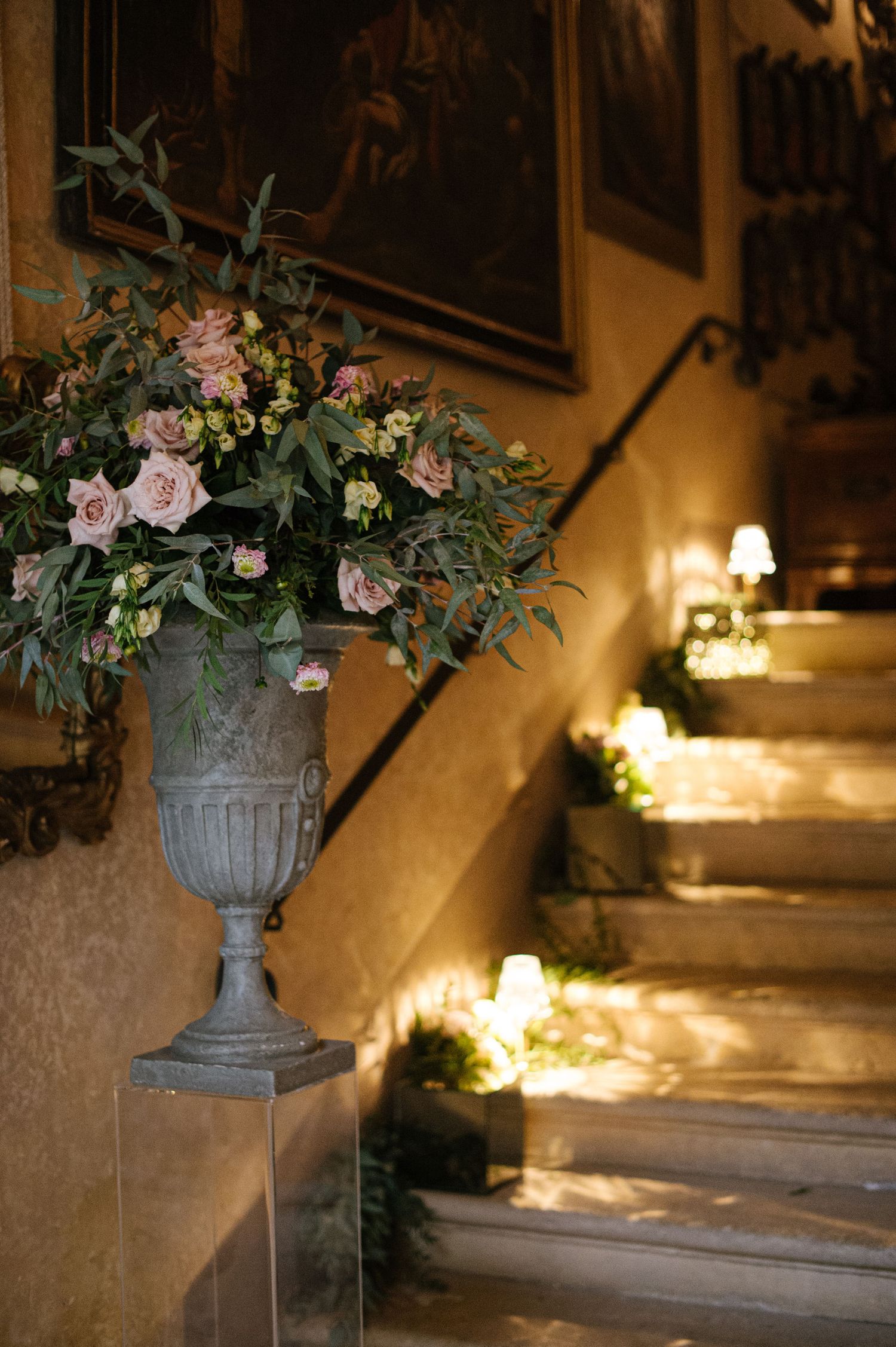 A classic stone urn filled with pink roses and eucalyptus sits beside candlelit stairs.