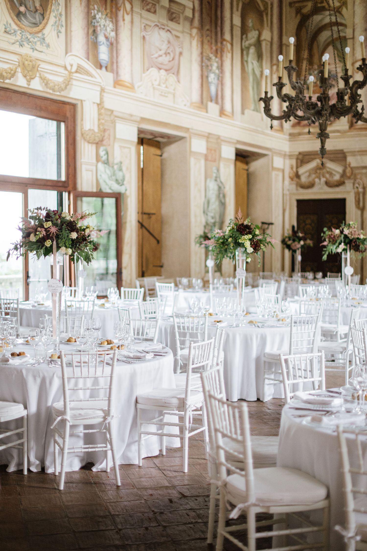 White wedding reception setup in grand ballroom with frescoed walls and crystal chandeliers.