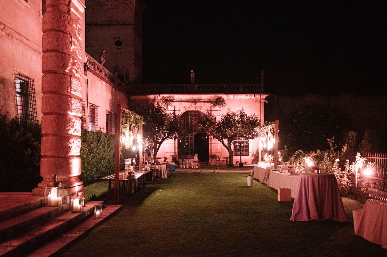 Outdoor evening wedding reception with pink lighting and cocktail tables along stone pathway.