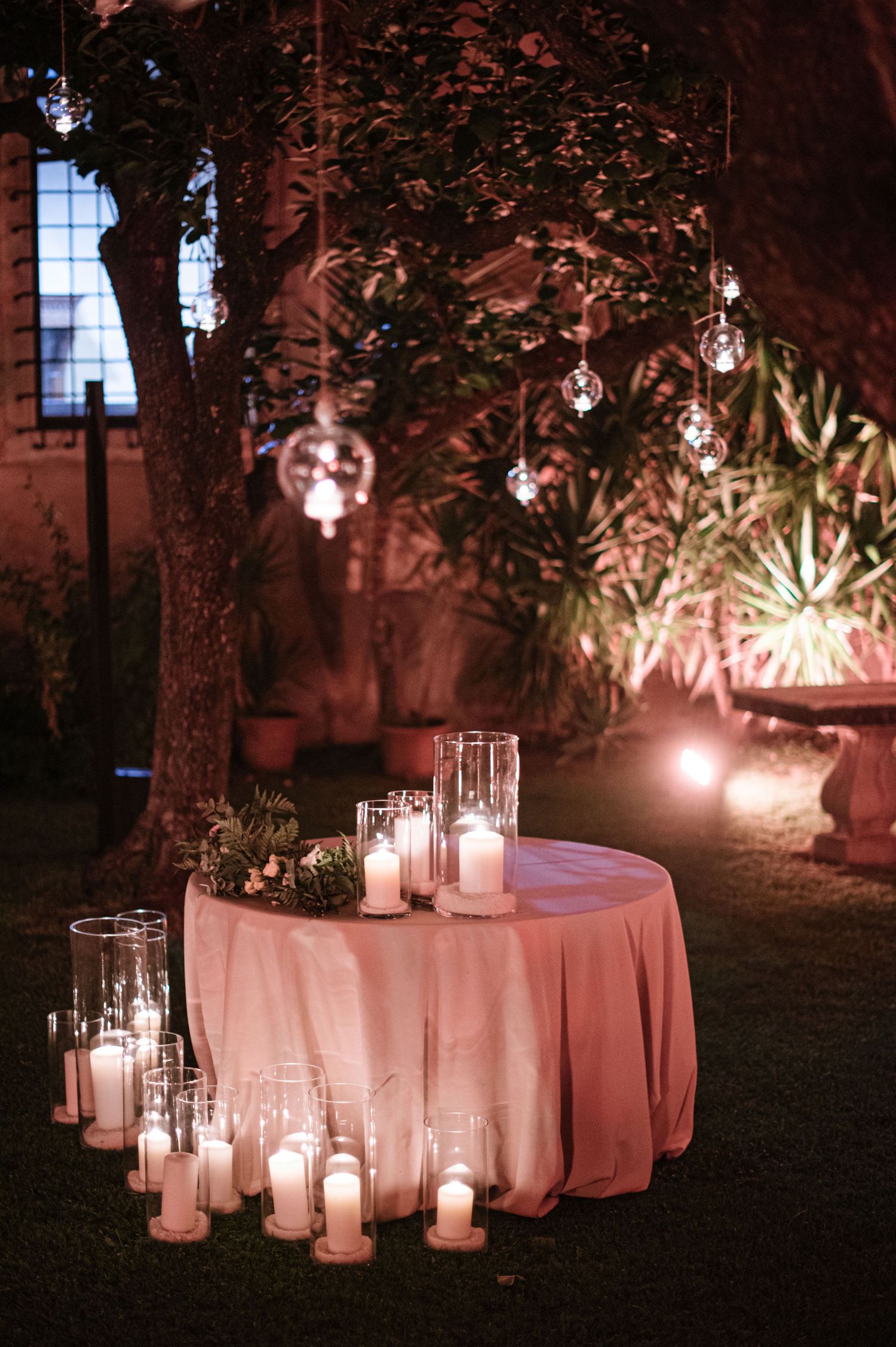 Romantic outdoor wedding decor with candles and pink tablecloth under hanging lights.