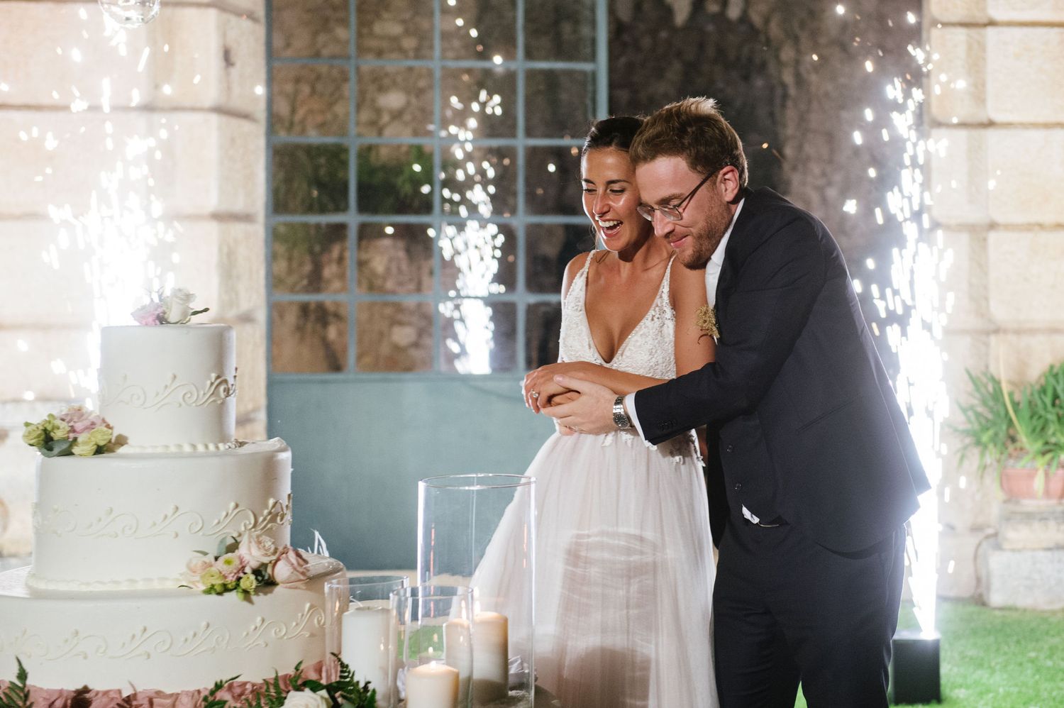 Wedding couple cutting cake with sparkler fountains in background at outdoor venue.