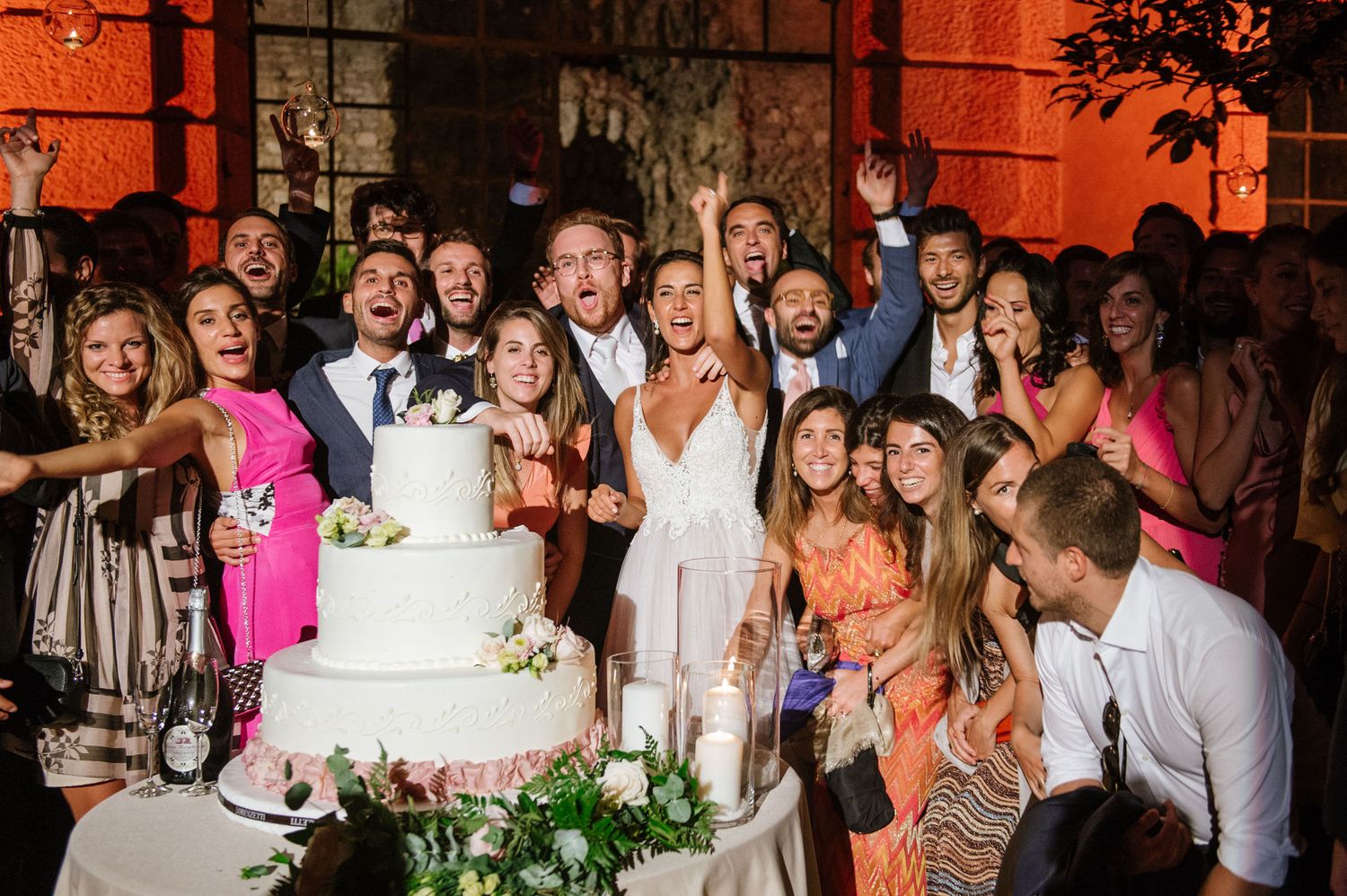 Wedding party and guests celebrating around tiered white cake at outdoor evening reception.