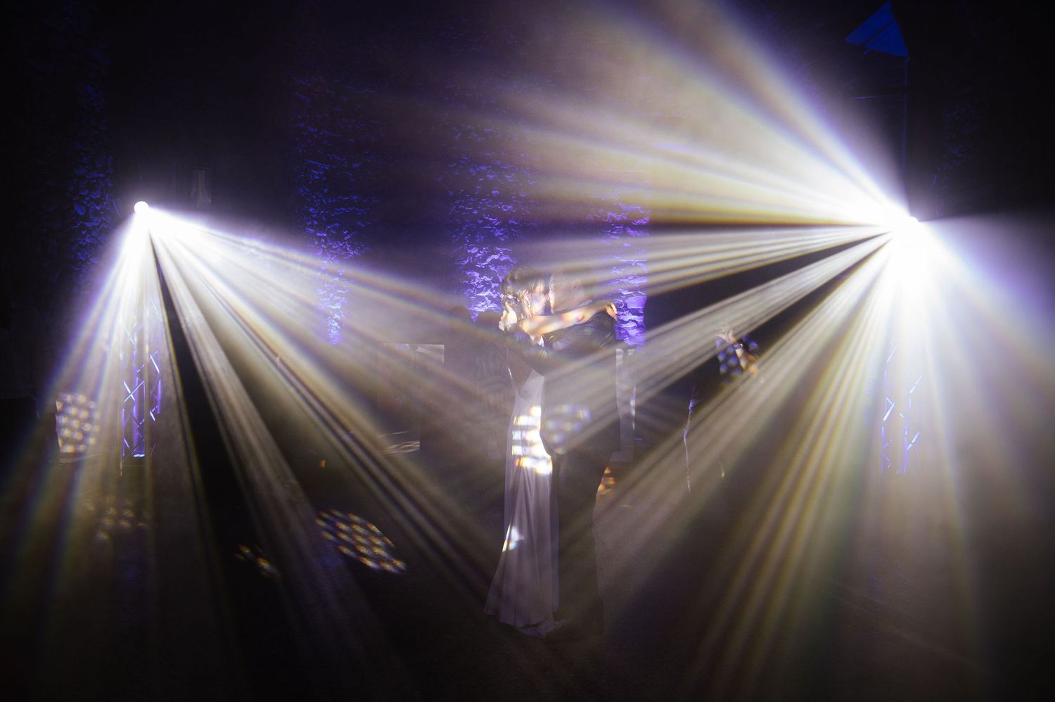 Multiple bright white spotlights create dramatic light beams in a dark reception venue.
