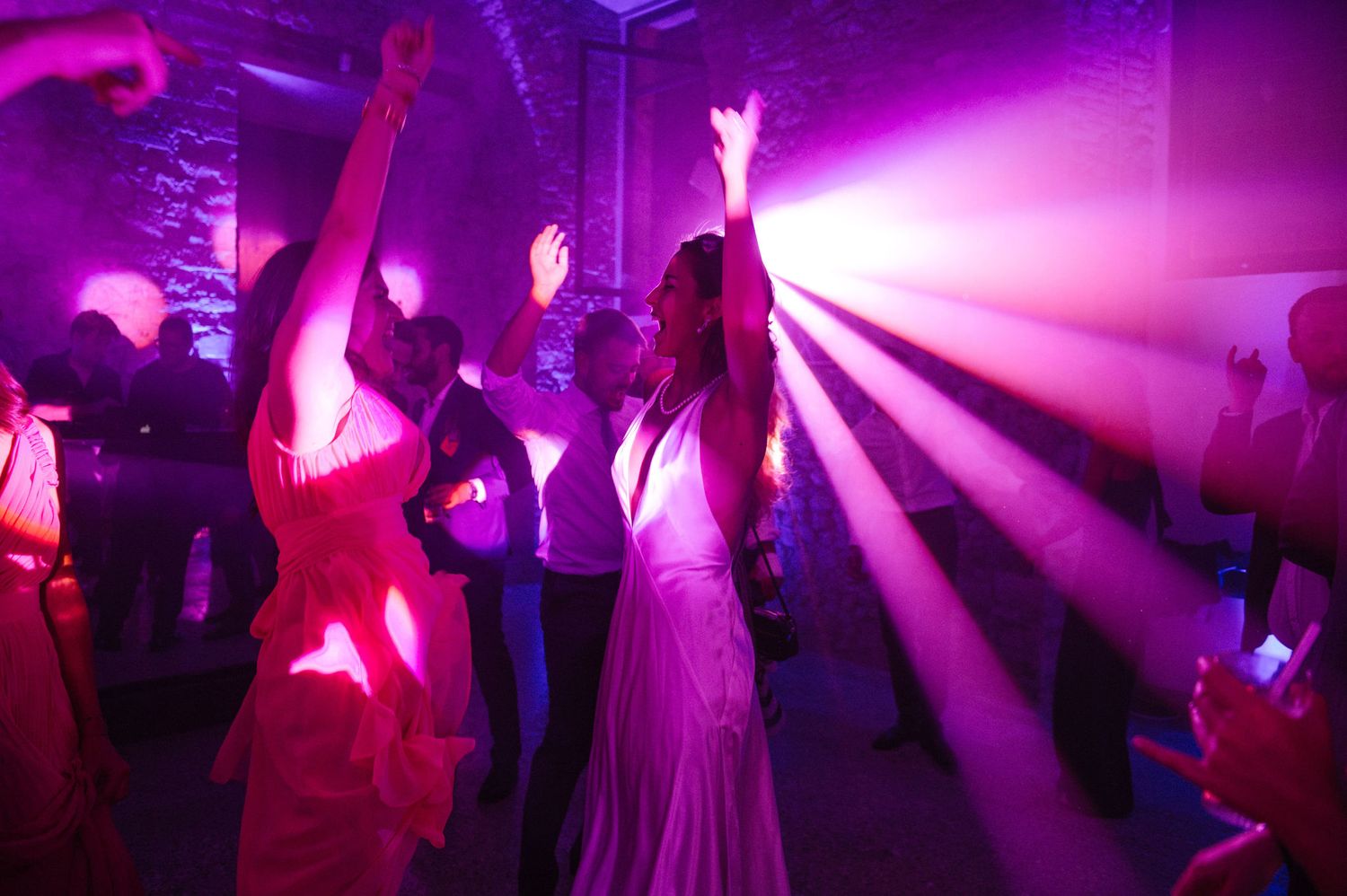 Wedding party dancing under purple spotlights and beams in a dimly lit venue.