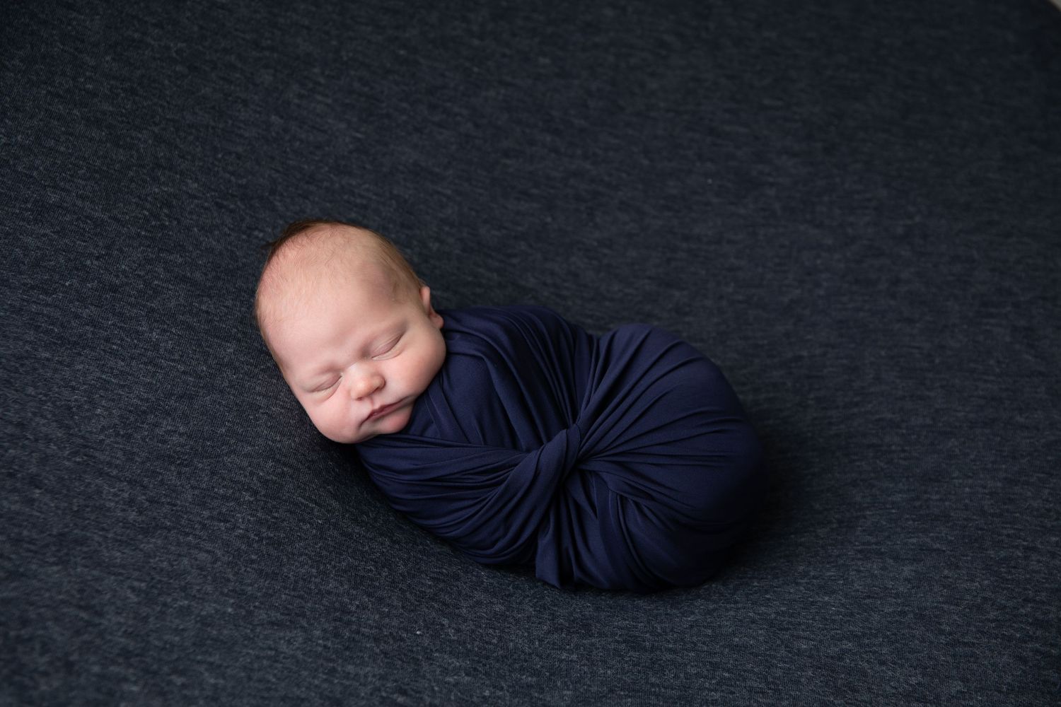 Sleeping newborn wrapped in navy blue fabric against dark background.