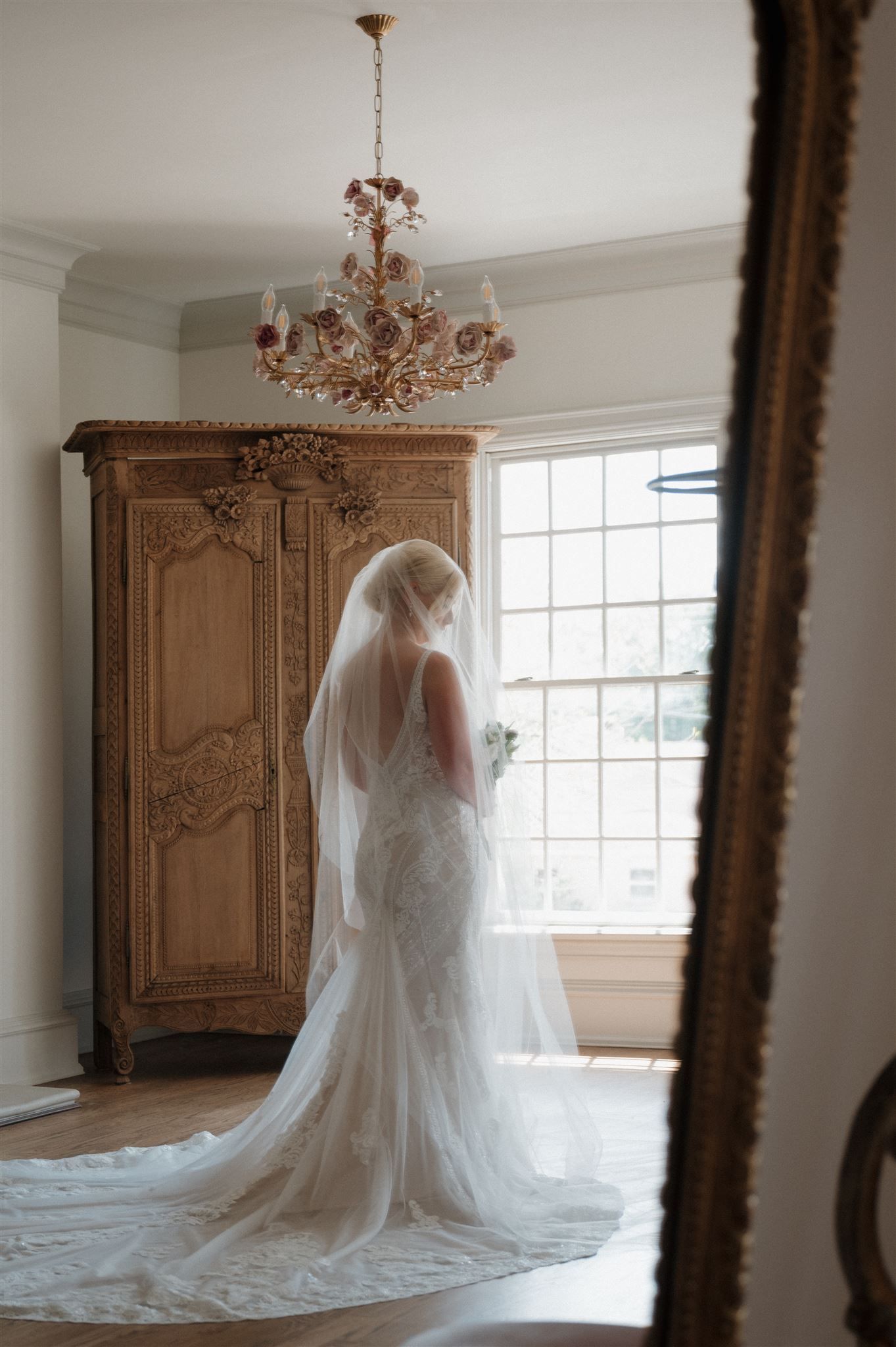 Ethereal silhouette of a bride in flowing veil against window light and vintage furniture.