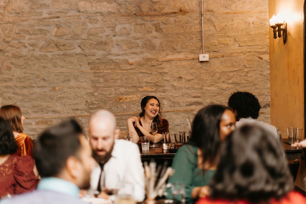 Woman smiling joyfully at a wedding reception dinner table in a stone-walled venue.