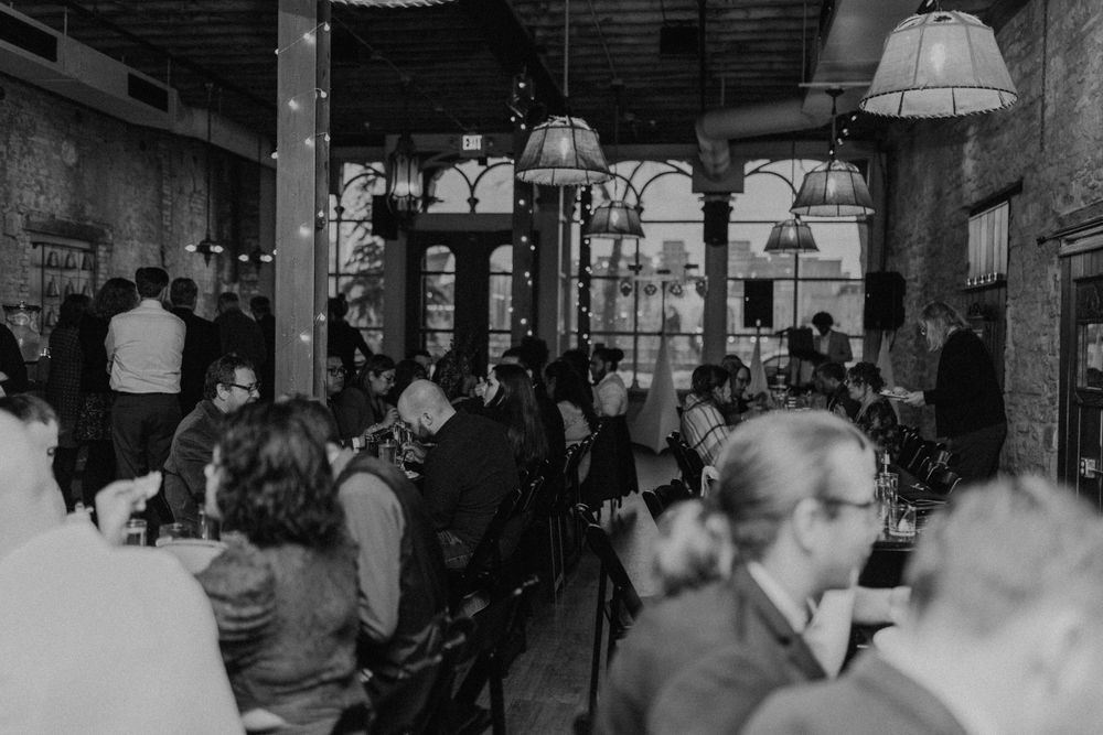 Black and white photo of wedding reception guests mingling in a warmly lit rustic brick venue.