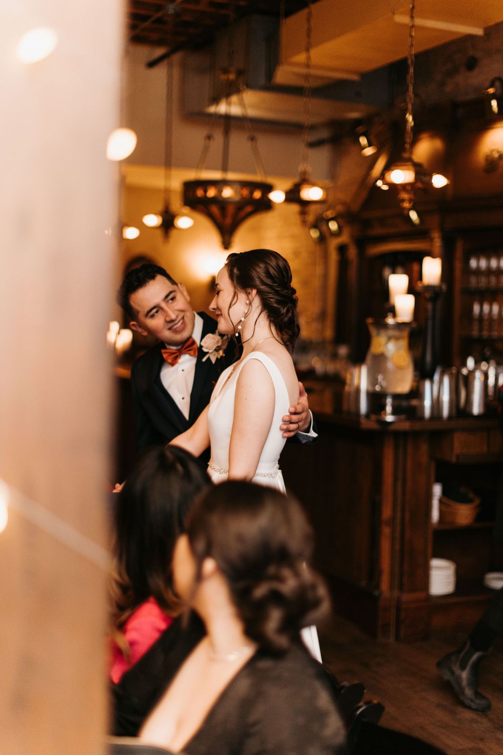 Groom in tuxedo and bride in white gown share a joyful moment on the warmly lit dance floor.