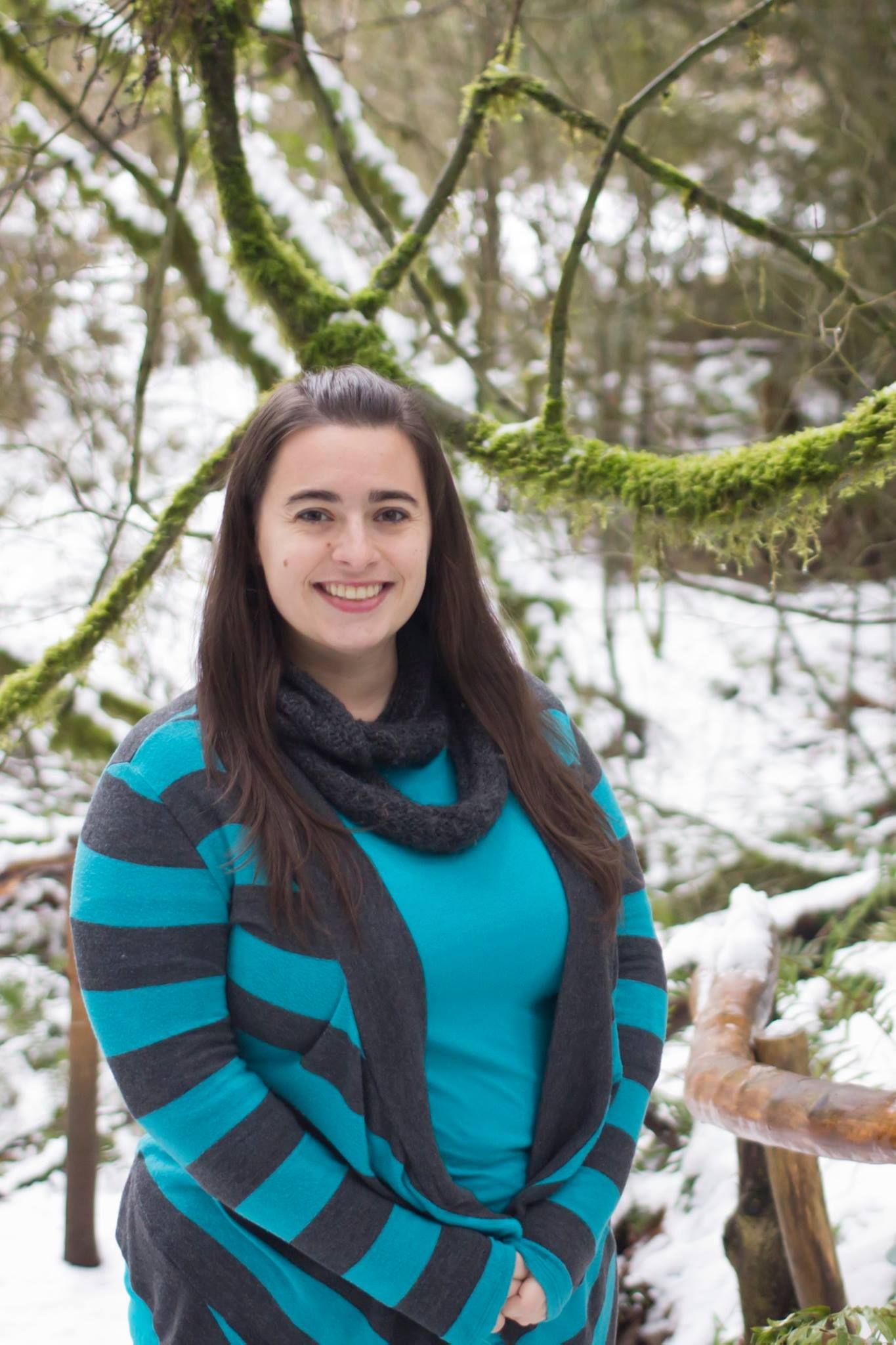 Person in turquoise and gray striped sweater standing in snowy forest with mossy trees.