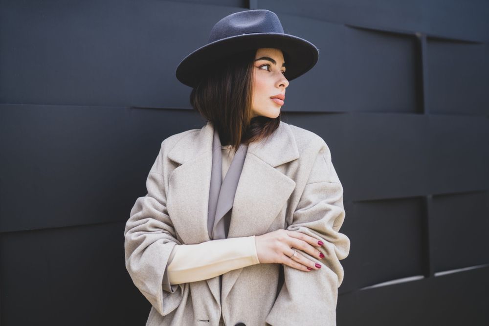 Stylish person in winter coat and wide brim hat poses thoughtfully against textured wall.