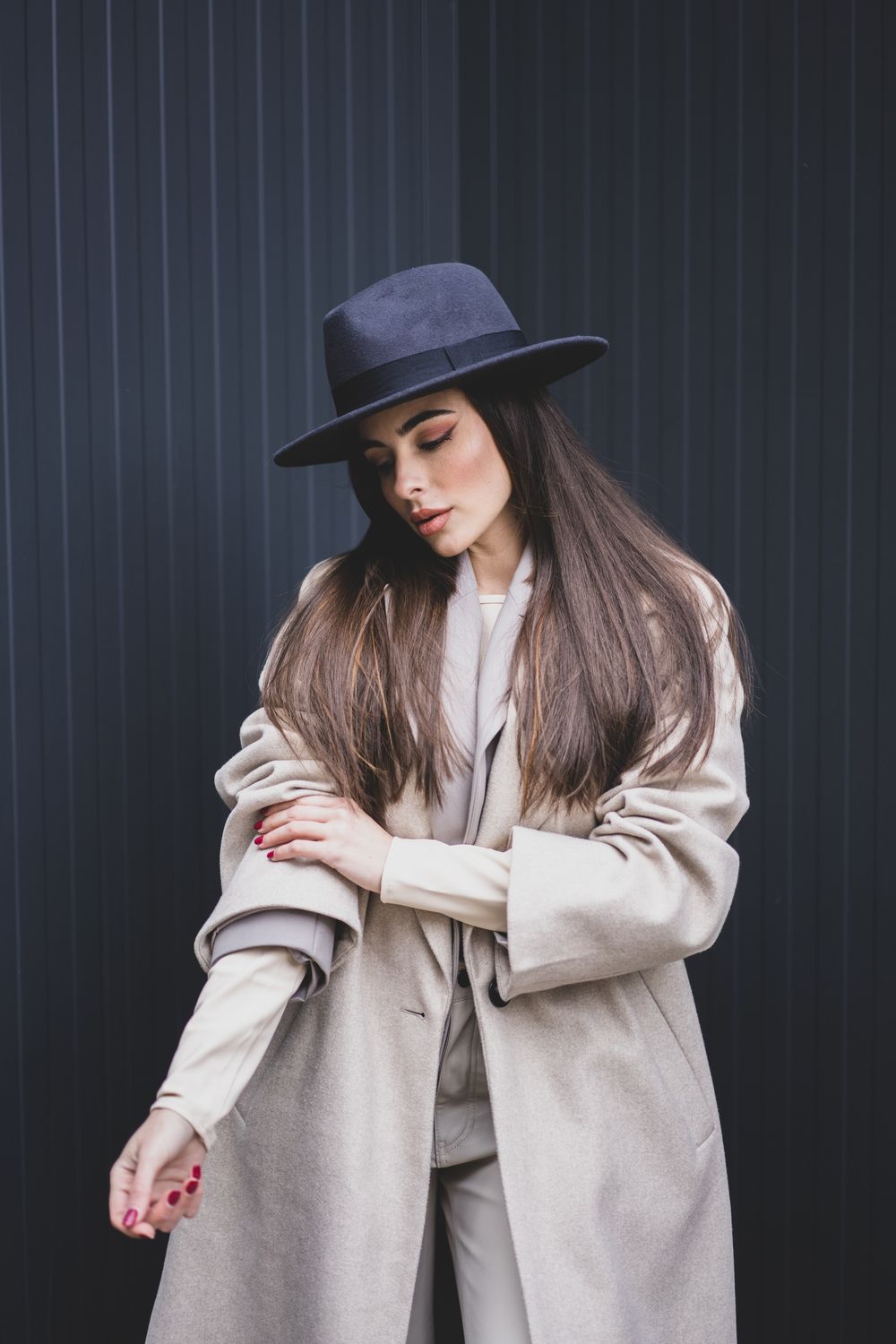 Fashion model in beige coat and navy hat poses against dark striped wall.