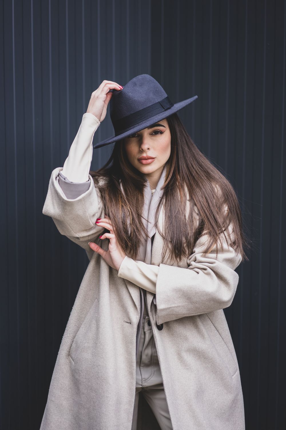 Stylish woman in neutral tones adjusts wide-brimmed hat against dark background.