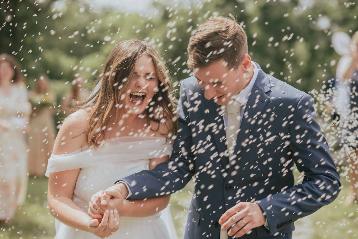 Newlywed couple laughs as guests shower them with white confetti in outdoor celebration.