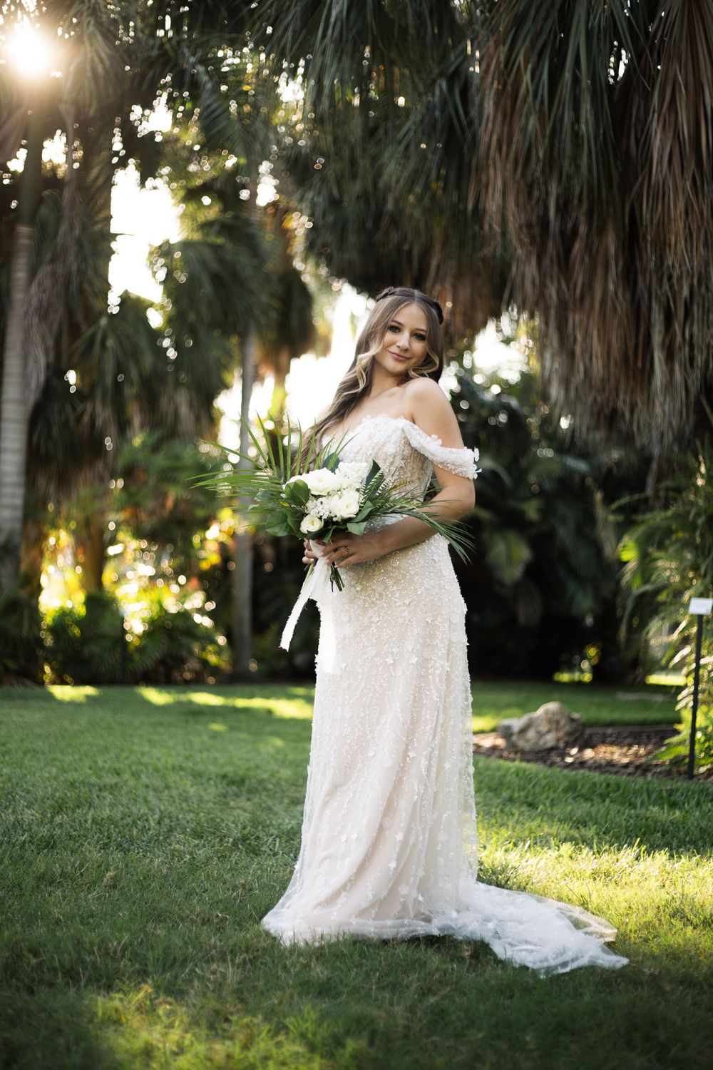 A stunning off-shoulder lace wedding dress stands out against a tropical garden backdrop at golden hour.