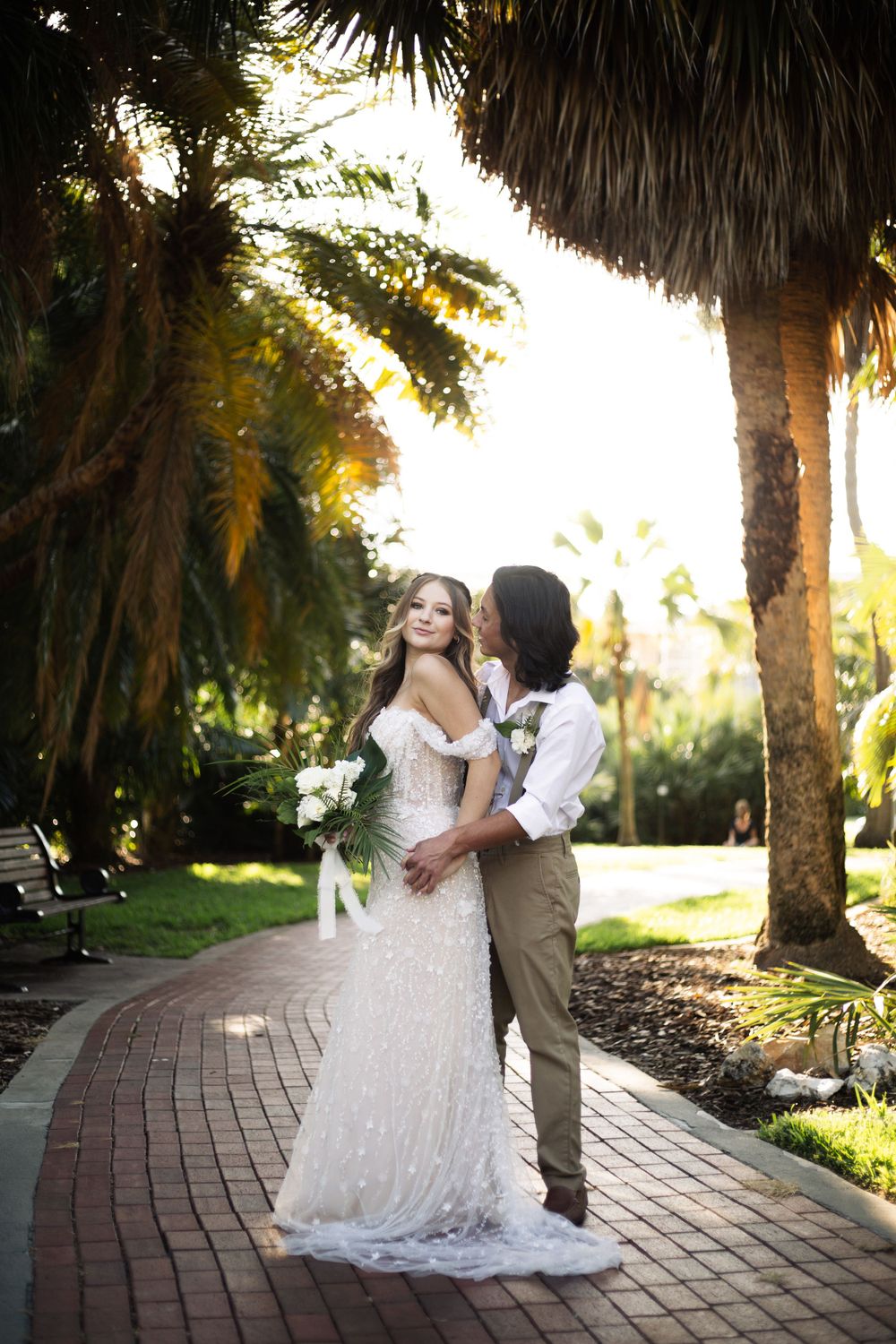 Newlyweds share an intimate moment on a winding brick path surrounded by tropical foliage.