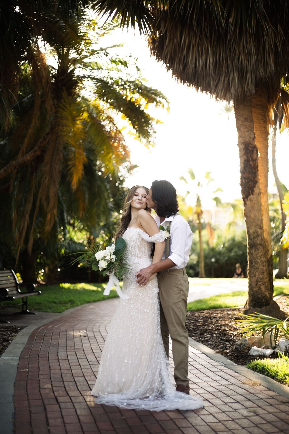 Romantic couple embracing on a brick pathway under palm trees during a wedding photoshoot.