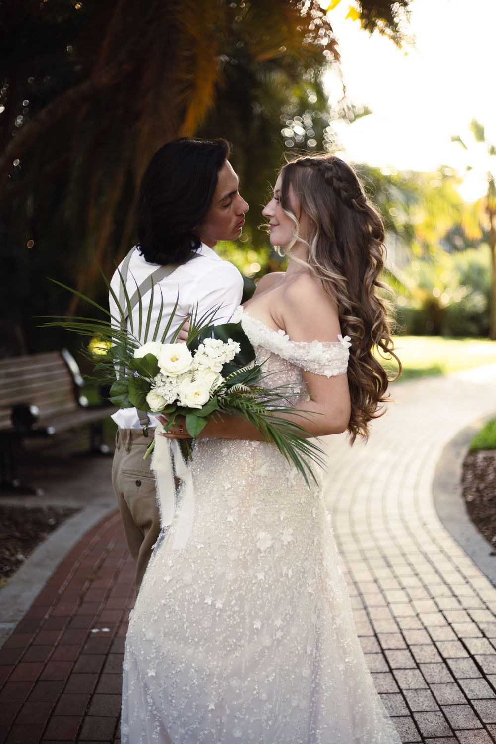 Newlyweds embrace on a brick path while holding an elegant white bouquet against a golden sunset backdrop.