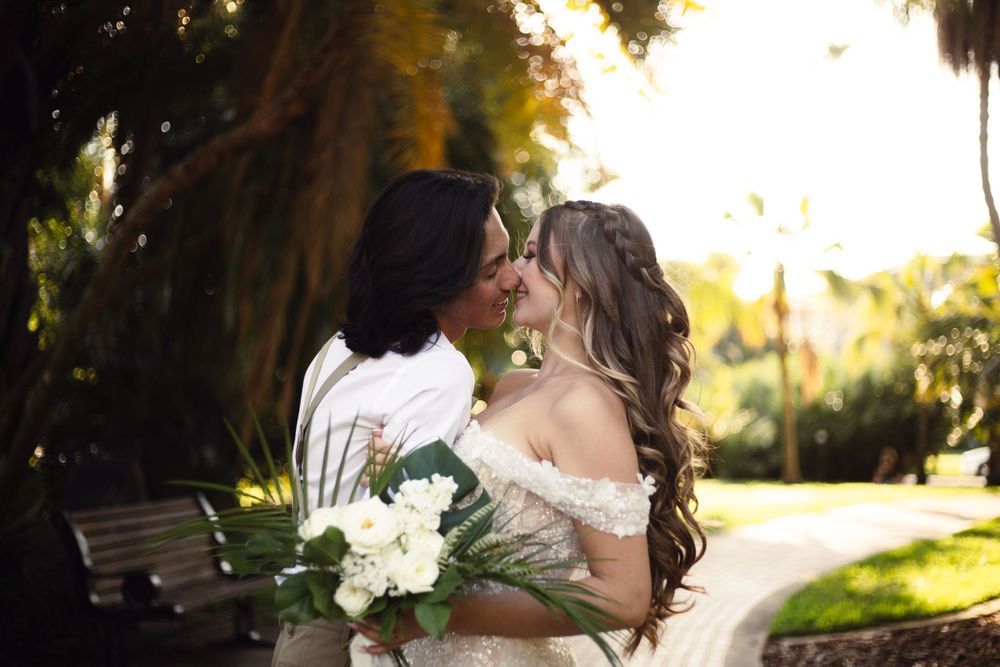 A romantic wedding couple sharing a tender moment in a garden setting with white floral bouquet at sunset.