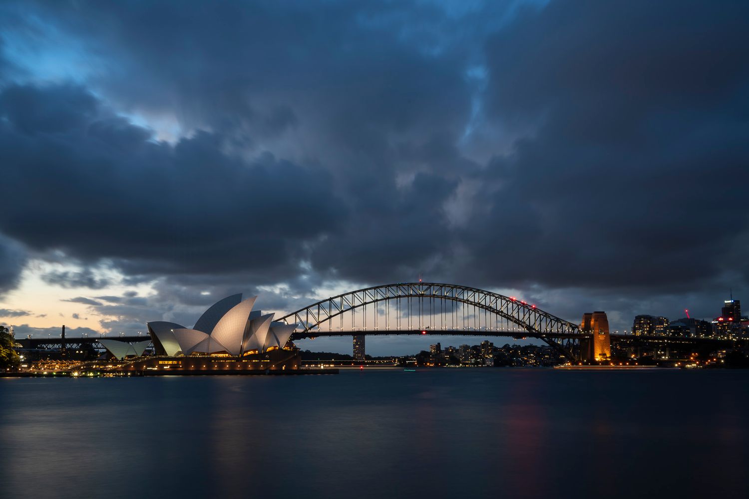Panoramic twilight view of Sydney Harbor Bridge and Opera House under dramatic storm clouds.