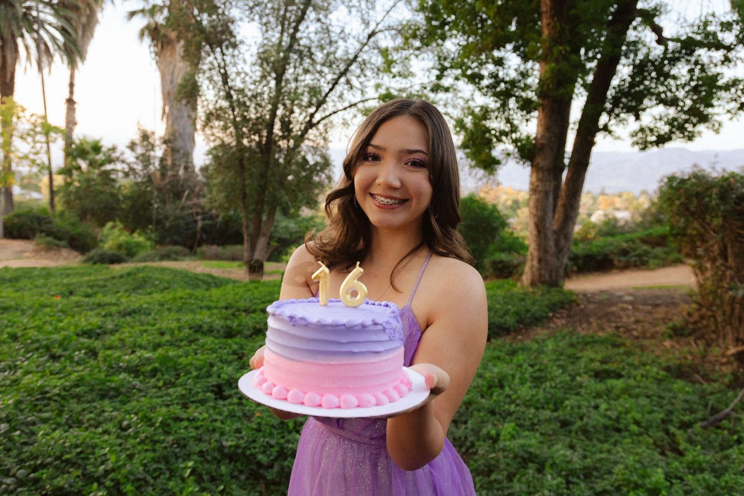 Person holding a pink birthday cake in an outdoor park setting with palm trees.