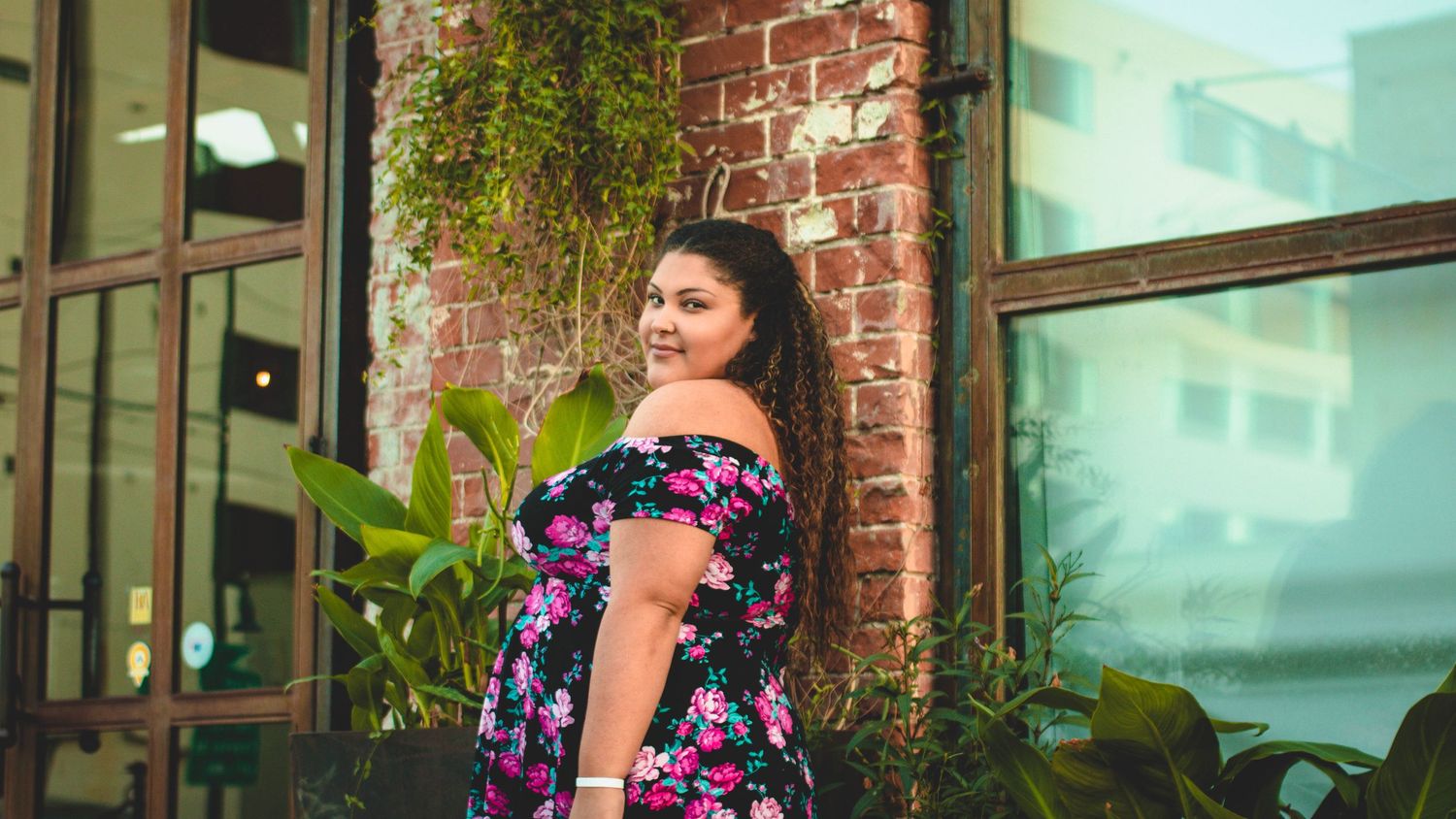 Person in floral dress poses near brick wall and hanging plants in urban setting.
