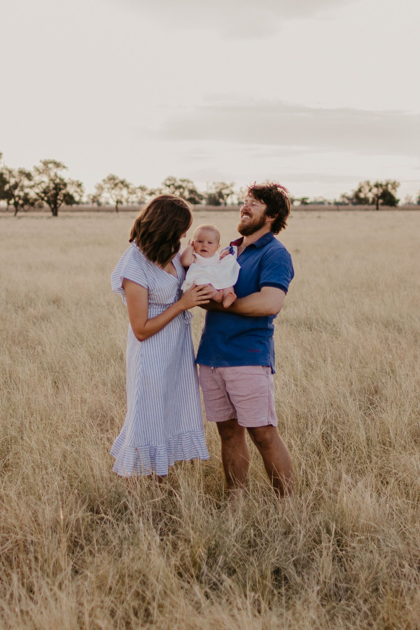 Baby held between mum and laughing dad, in a long grass paddock with fence and trees in the background