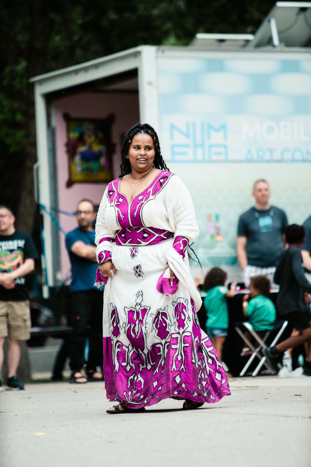 A person wearing a flowing white and pink patterned dress stands in front of a white trailer.