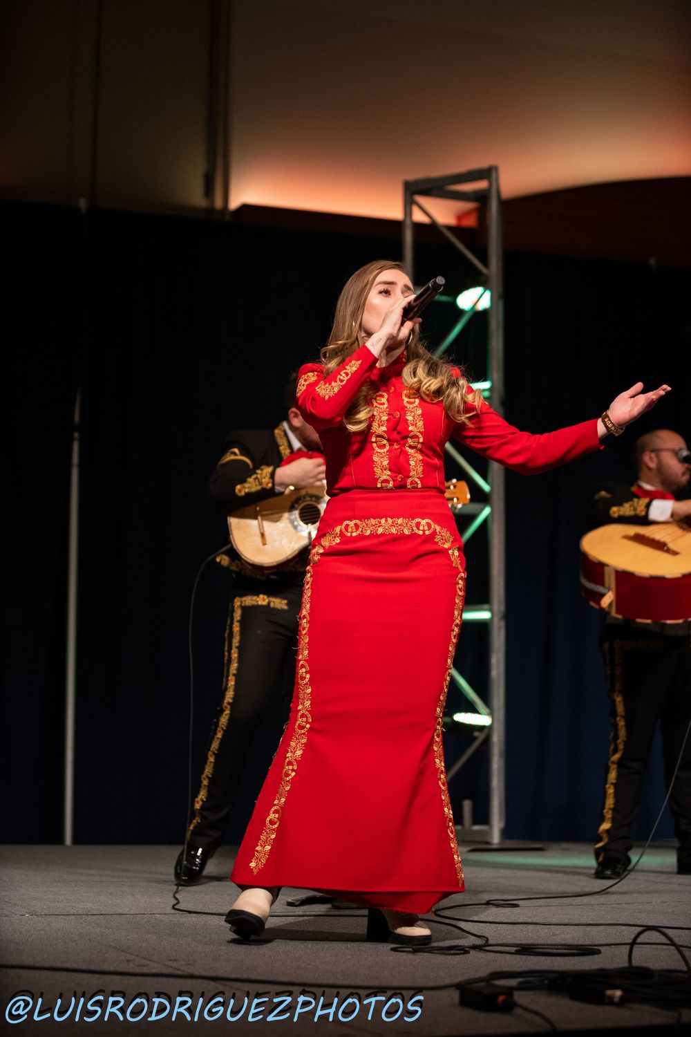 A performer in a red and gold traditional dress singing on stage during a cultural performance.
