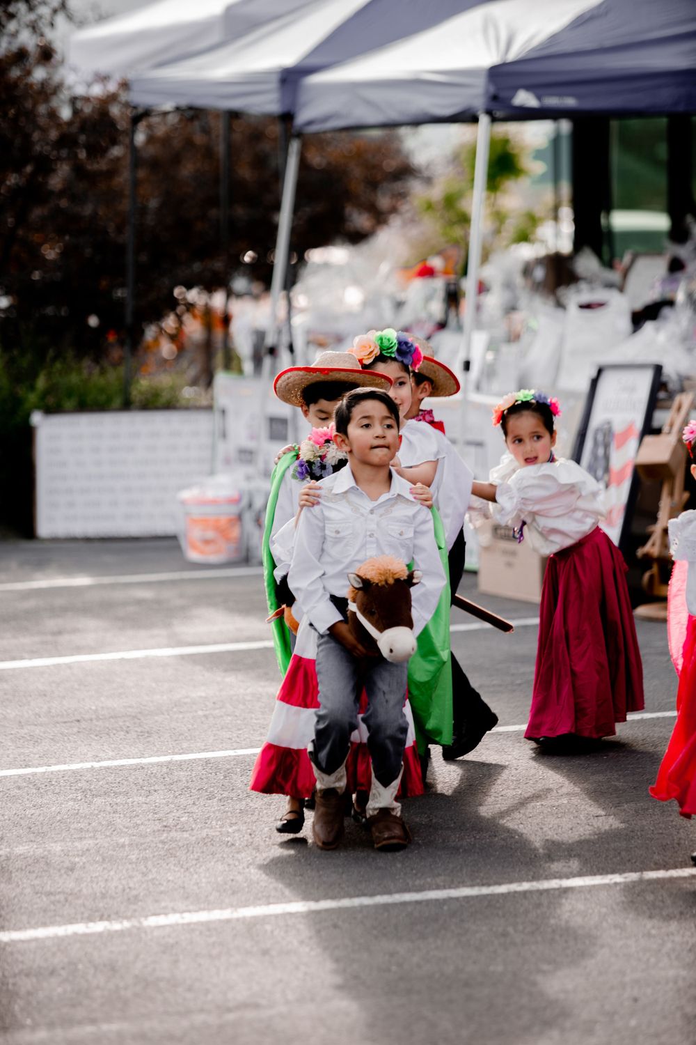 Young dancers perform traditional Mexican folk dance in colorful costumes under white canopies in a parking lot.