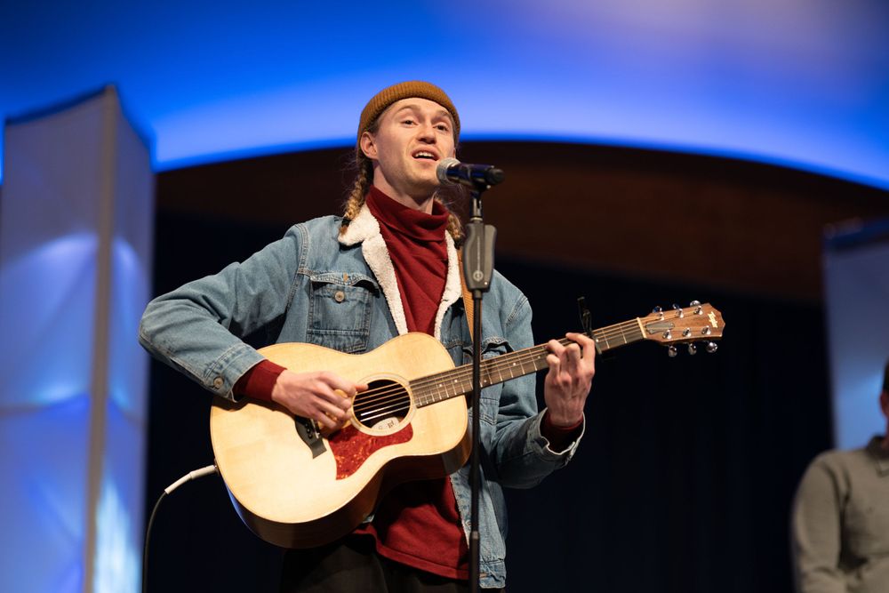 Musician in denim jacket and red beanie performs with acoustic guitar on stage.