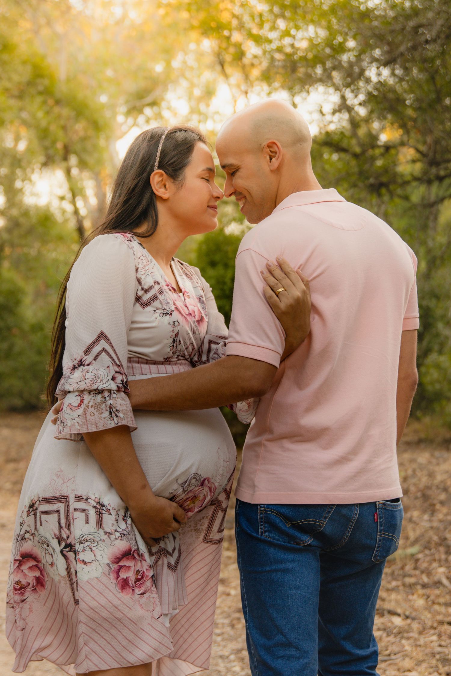 A romantic maternity photoshoot in a park with soft golden sunlight filtering through trees.